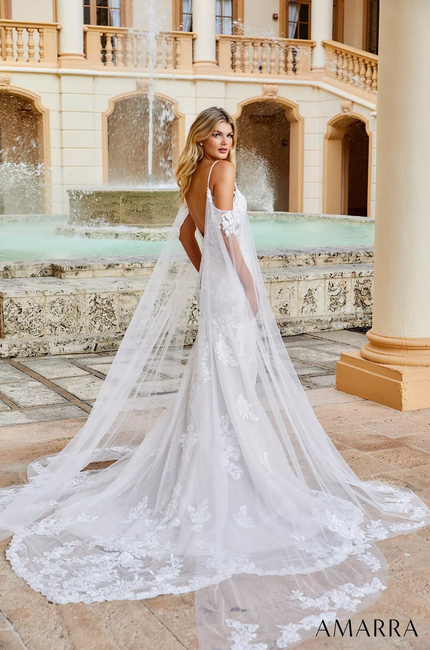 A bride in a white lace gown with flowing cape poses by a fountain in an elegant Mediterranean-style courtyard.