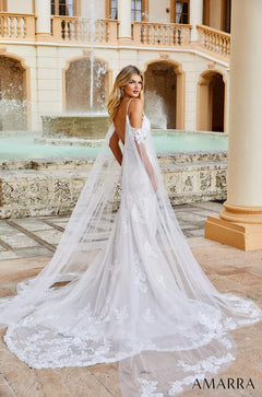 A bride in a white lace gown with flowing cape poses by a fountain in an elegant Mediterranean-style courtyard.