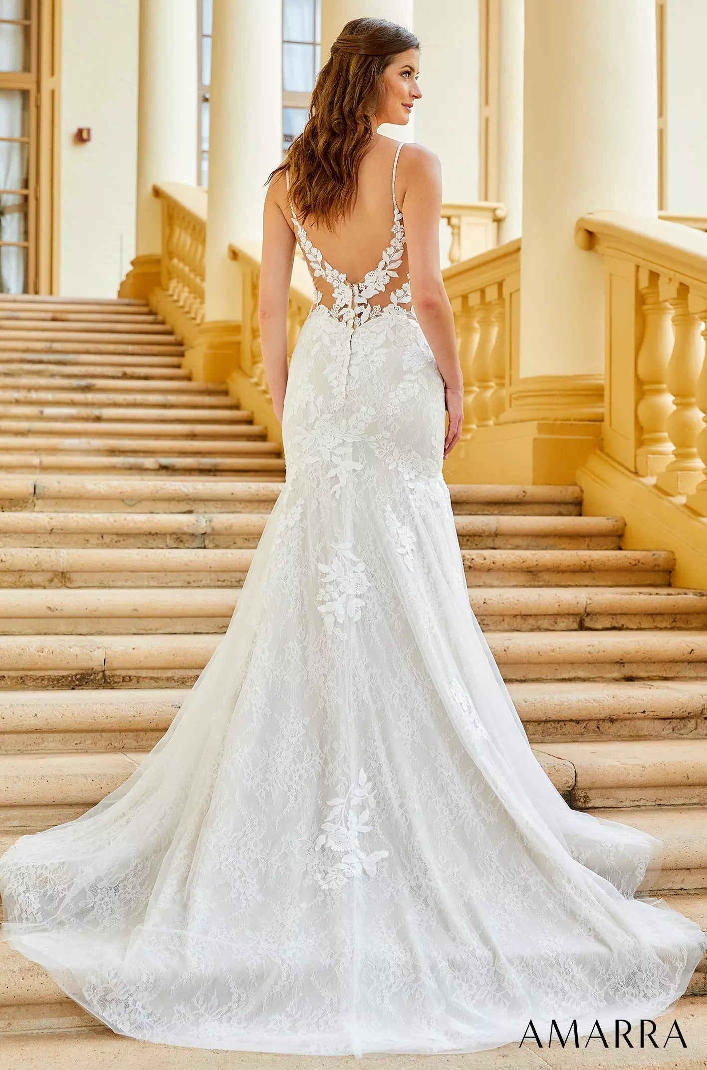 A bride in a white lace mermaid wedding dress with a flowing train stands on marble stairs.