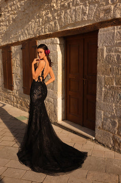 A woman in a black lace mermaid gown with a long train stands against a stone wall at sunset.