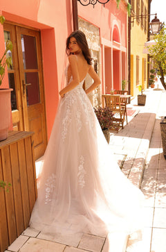 A bride in a delicate lace wedding gown stands on a charming Mediterranean street with pastel buildings.