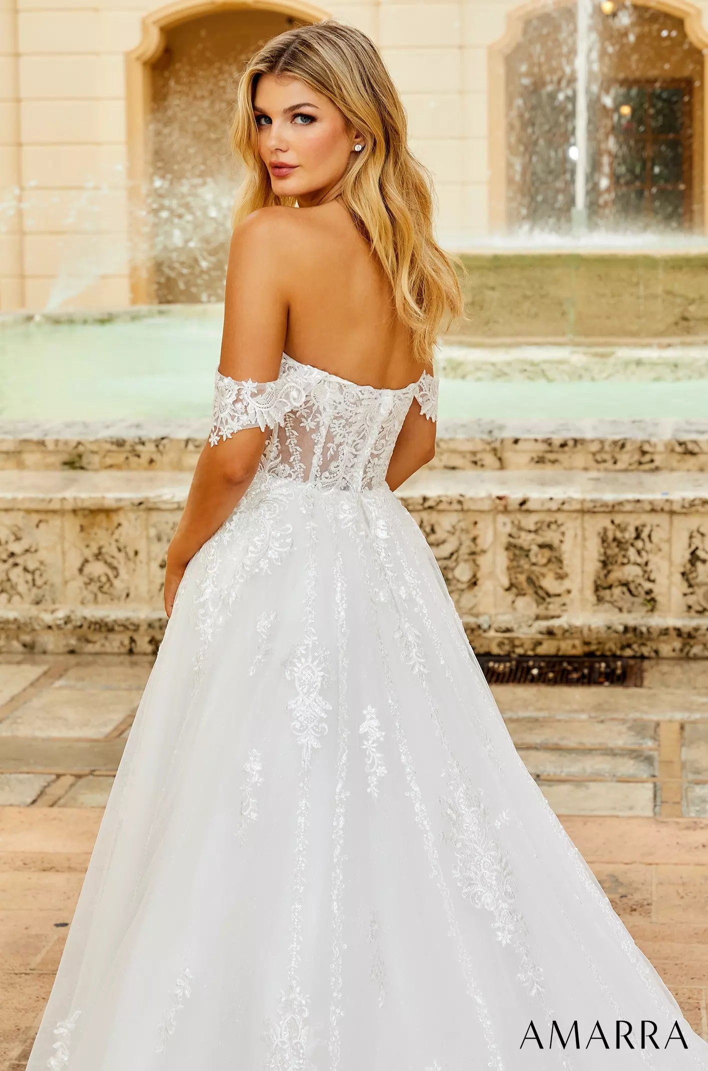 A blonde model wears an off-shoulder white lace wedding gown with a full skirt, standing near a fountain.