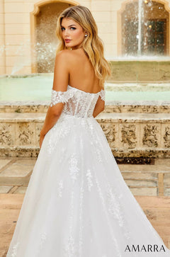 A blonde model wears an off-shoulder white lace wedding gown with a full skirt, standing near a fountain.