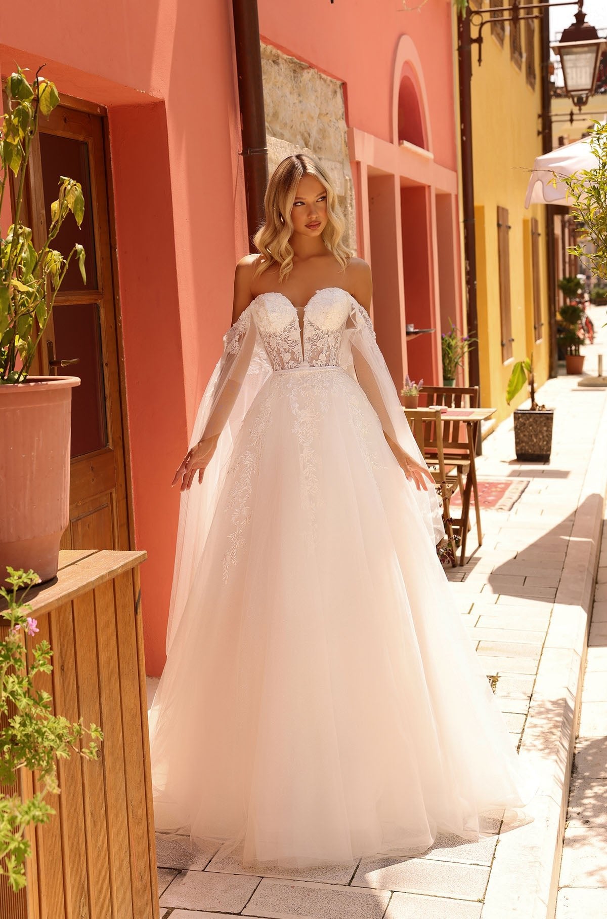 Bride in off-shoulder lace ballgown stands on charming cobblestone street with pastel buildings.