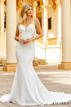 A blonde woman wears an elegant white mermaid-style wedding dress with lace details against a classical columned background.