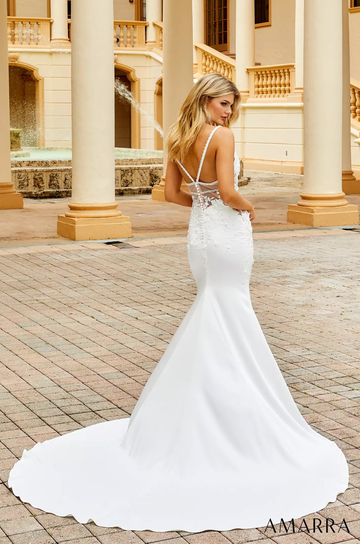 A blonde woman wears a white mermaid-style wedding gown with a sheer lace back against an ornate columned building.