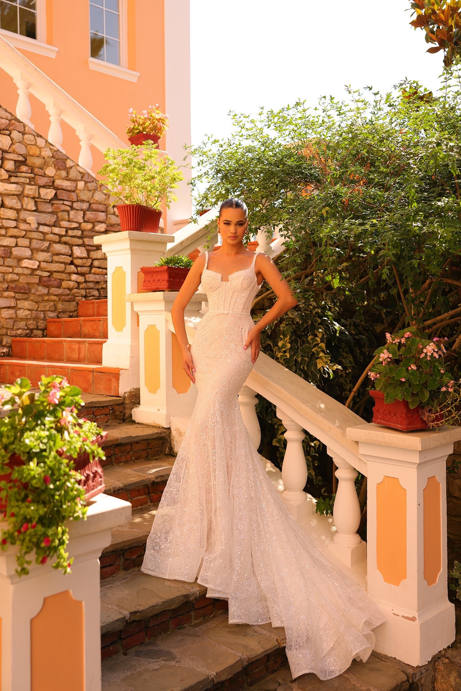 A bride in a sparkling white mermaid wedding gown stands on stone steps with lush garden surroundings.