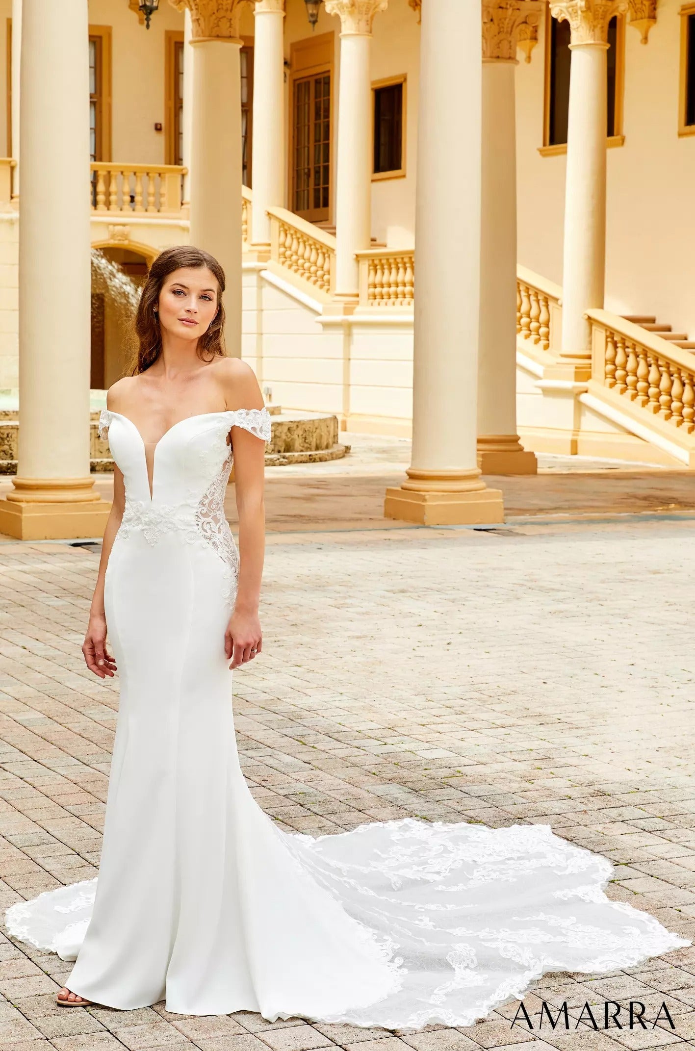 A bride in an off-shoulder white mermaid wedding gown stands on a columned terrace with a long lace train.