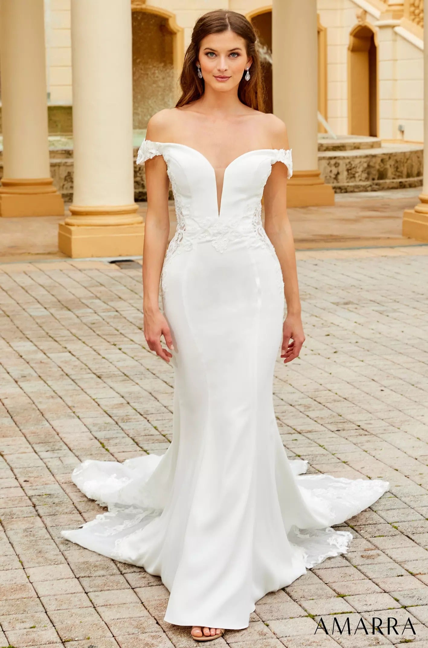 A white off-shoulder mermaid wedding dress with lace details, trailing a long train on a tiled outdoor patio.