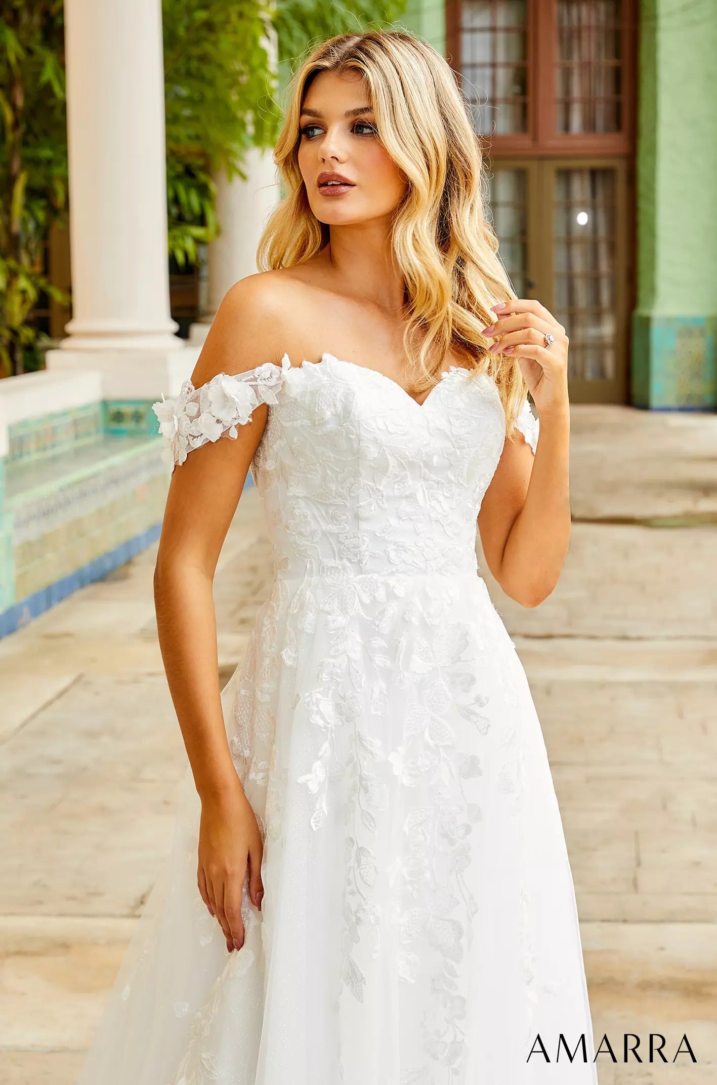 A blonde woman in an off-shoulder white lace wedding dress stands near white columns in soft, wavy light.