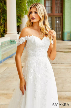 A blonde woman in an off-shoulder white lace wedding dress stands near white columns in soft, wavy light.