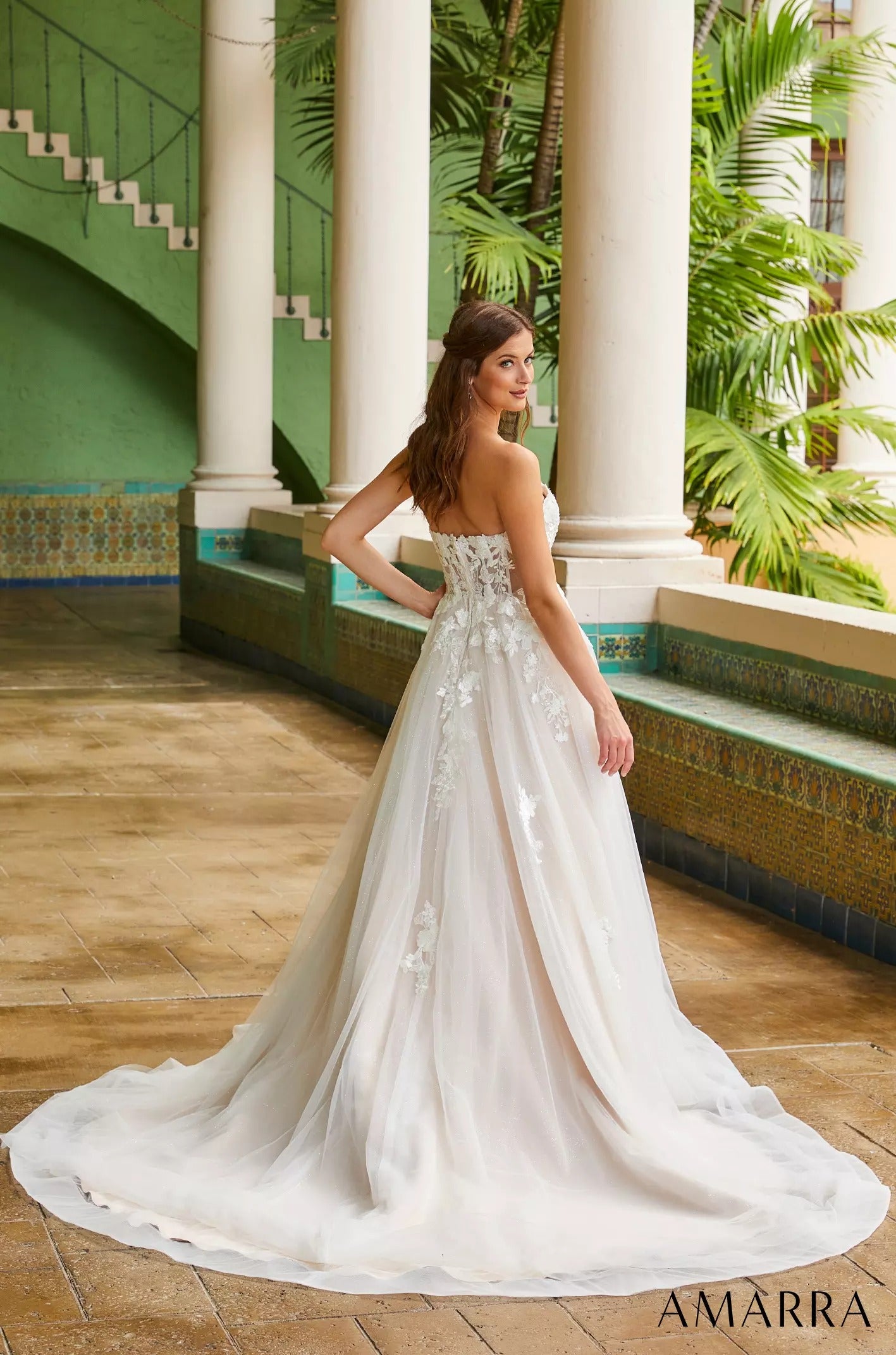 A bride in a strapless white wedding gown with floral details stands in a green-and-white columned interior.