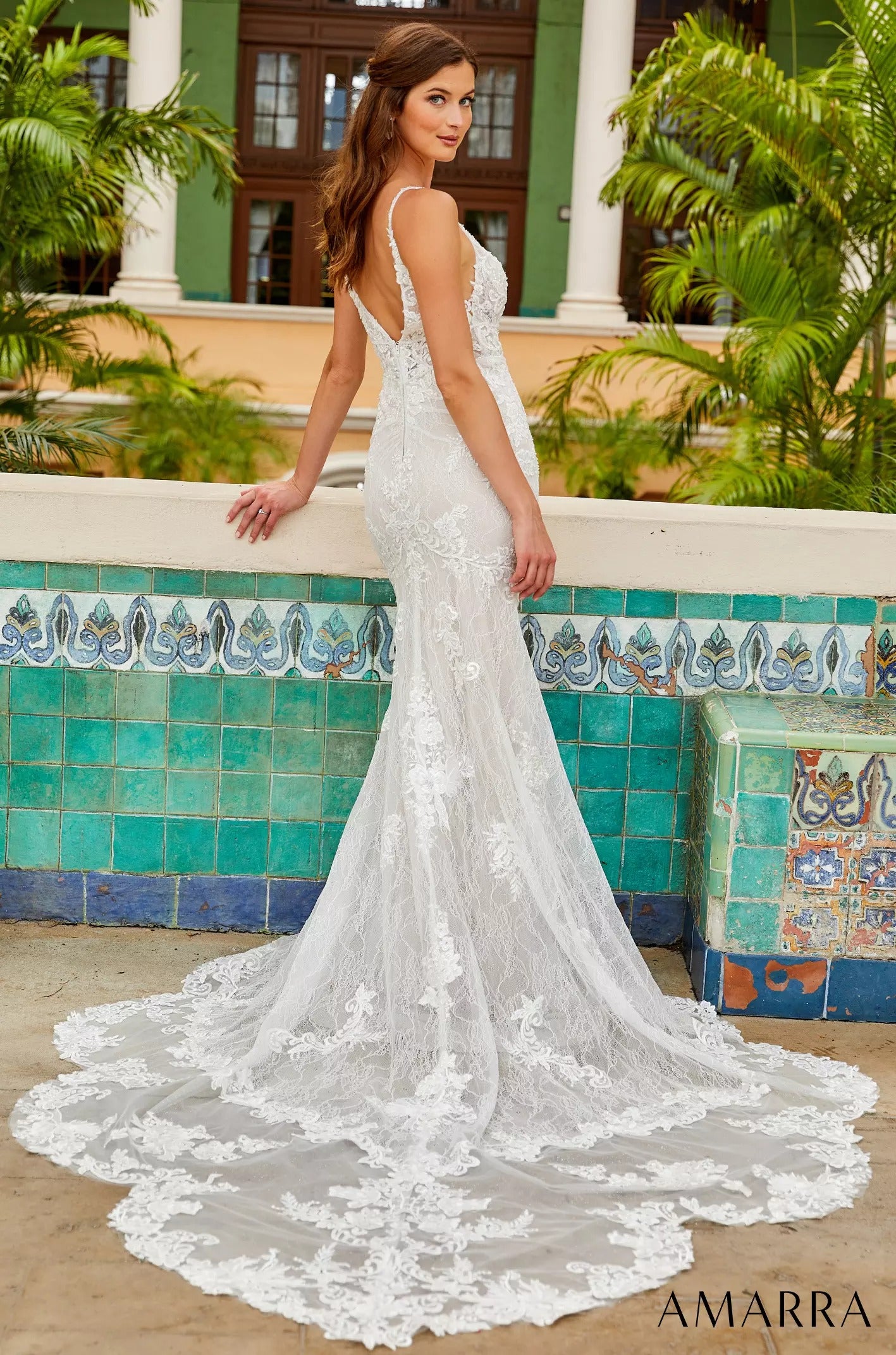 A white lace mermaid wedding dress with a long train, worn against a tropical background with green tiles.