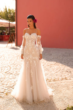 A white lace wedding gown with off-shoulder sleeves and floral appliqués, worn against a terracotta wall backdrop.