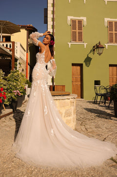A bride in a lace mermaid wedding dress poses on a cobblestone street with a green building behind her.