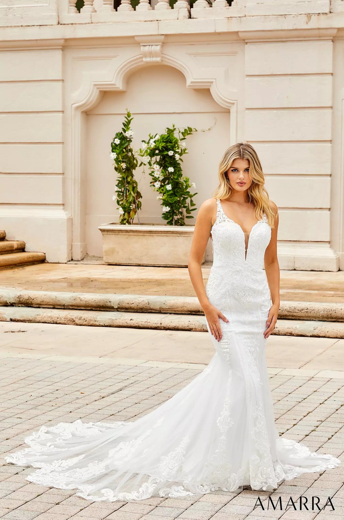 A blonde woman in a white lace mermaid wedding dress stands on stone steps near a white architectural backdrop.