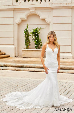 A blonde woman in a white lace mermaid wedding dress stands on stone steps near a white architectural backdrop.