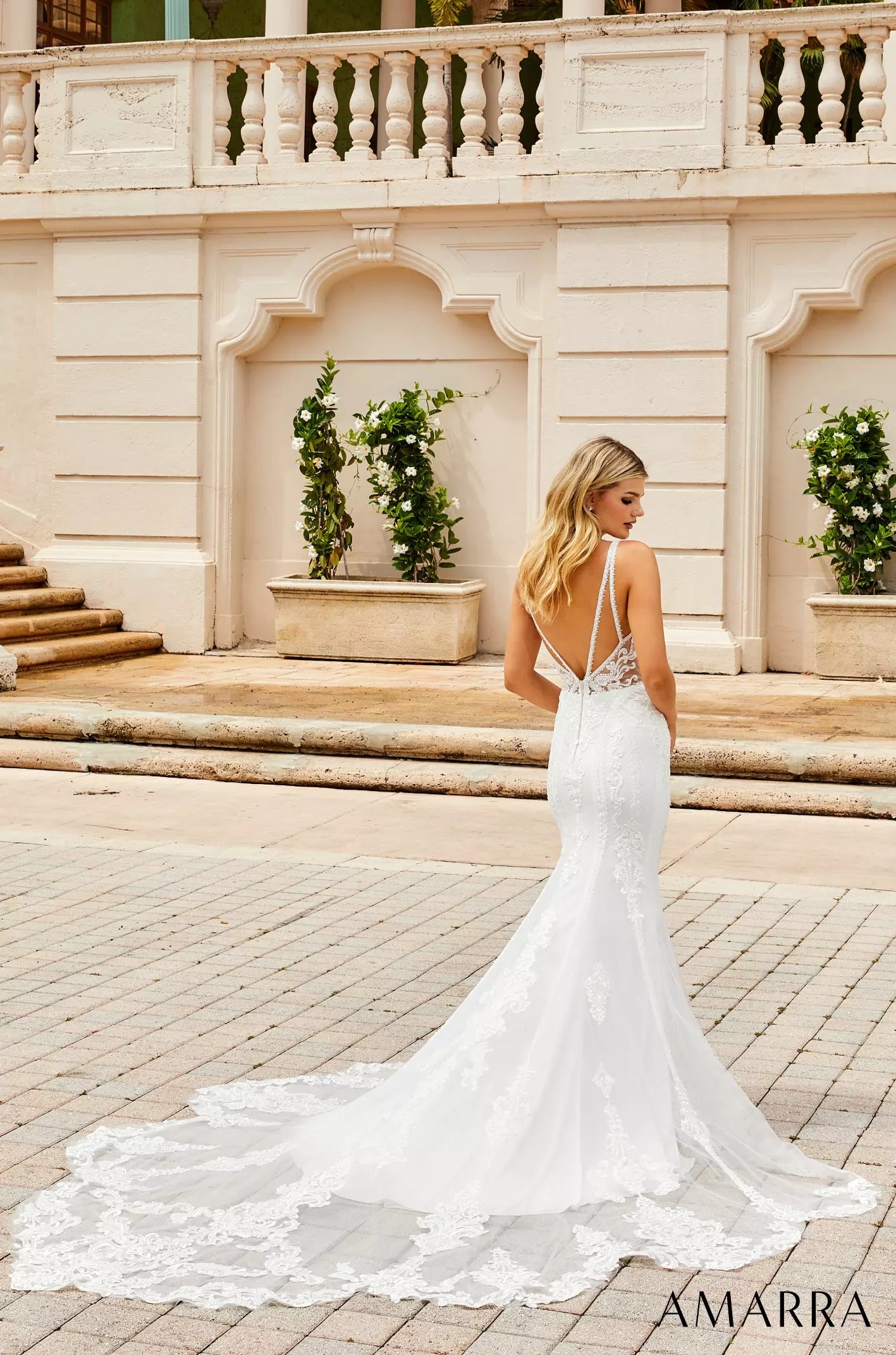 A bride in a white lace mermaid wedding dress stands on stone steps, facing away to show her low back and train.